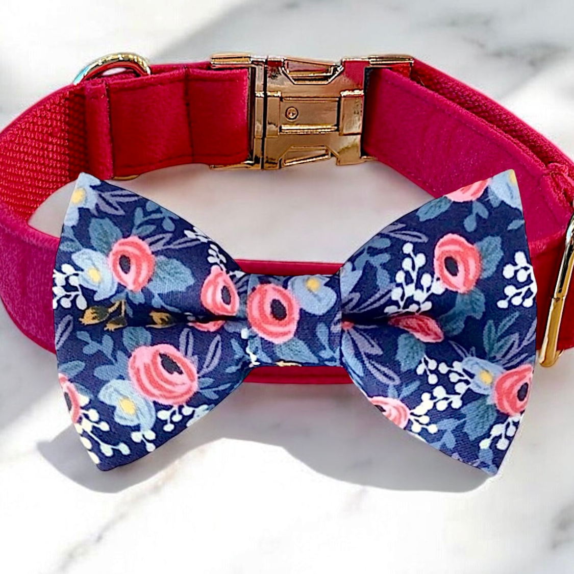 A pink collar with a gold buckle and a blue bow tie with a floral pattern are displayed on a white marble surface.
