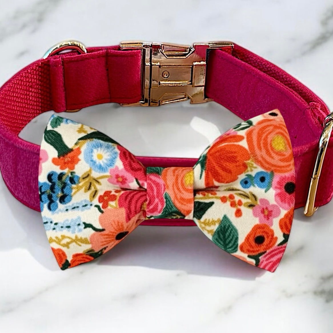 A pink collar with a gold buckle and a colorful bow tie with a floral pattern are displayed on a marble surface.