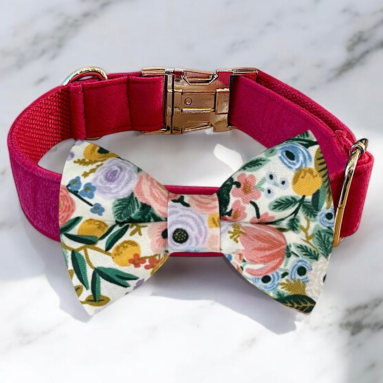 A red collar with a gold buckle and a bow tie with a floral pattern are displayed on a marble surface.