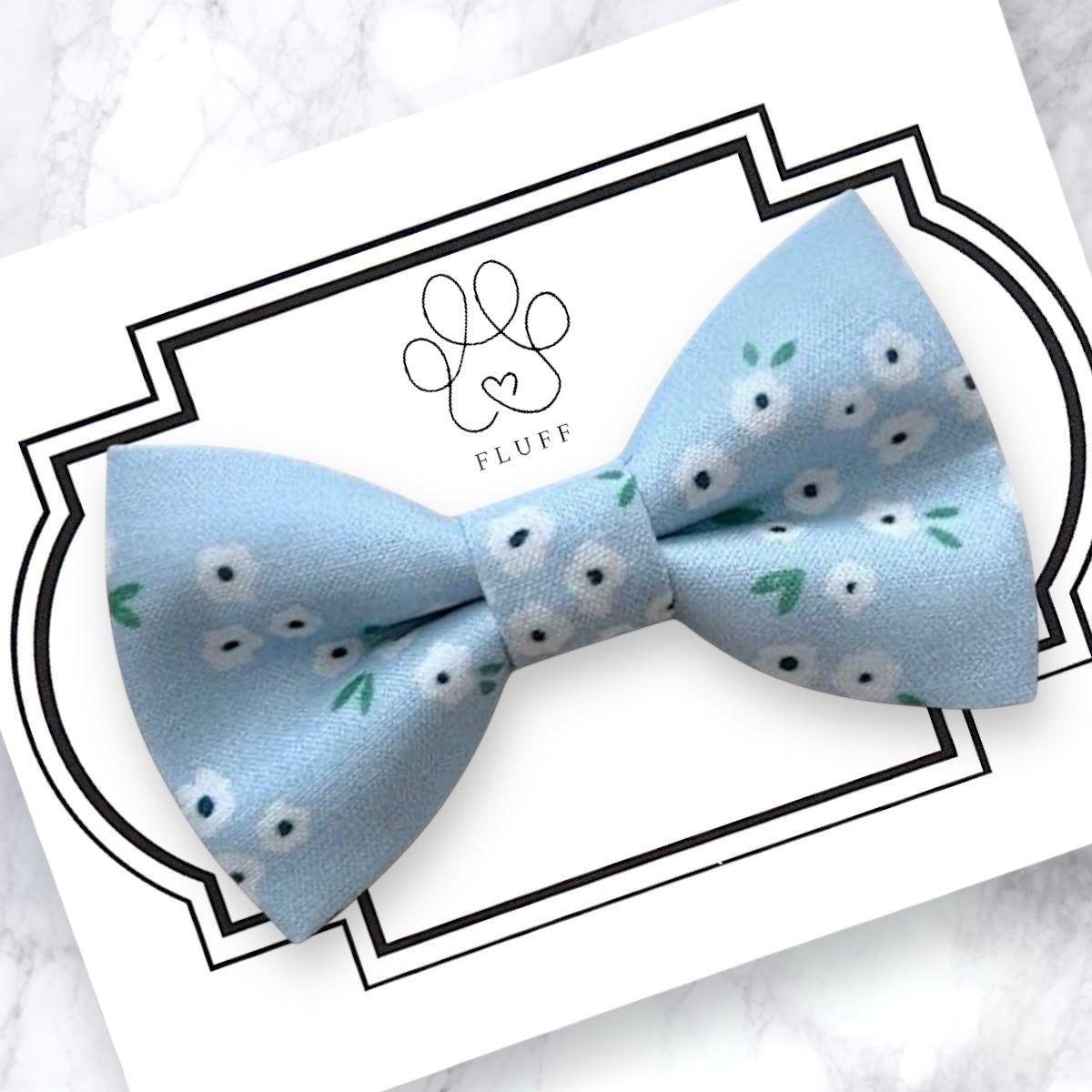 Light blue dog bow tie with floral pattern on a white card with 'Fluff' branding.