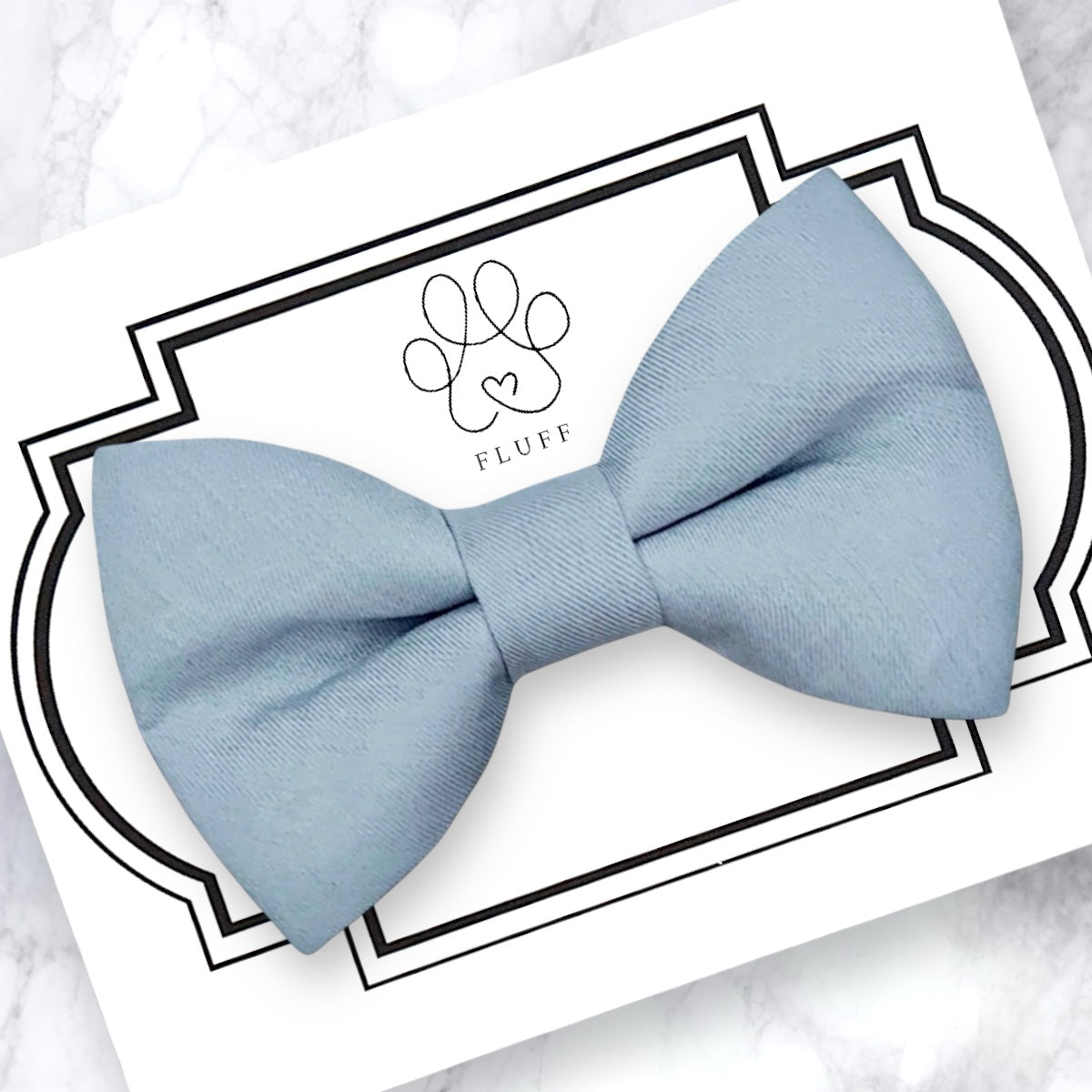 Dusty blue bow tie on a white card with a decorative border and brand logo.