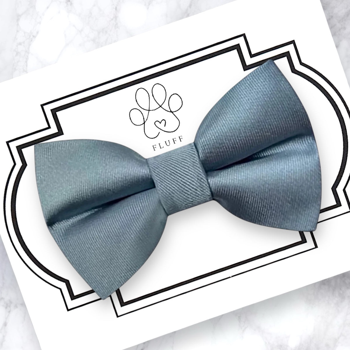 Dusty Blue dog bow tie on a white card with a brand logo, on a marble surface.