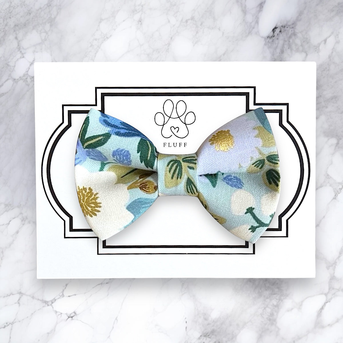 Blue Floral-patterned dog bow tie on a white card with 'FLUFF' branding on a marble background