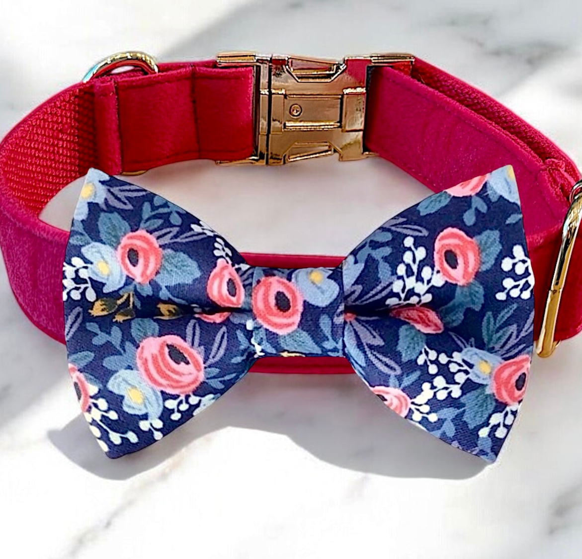 A pink collar with a gold buckle and a blue bow tie with a floral pattern are displayed on a white marble surface.