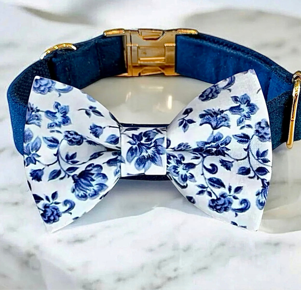 Navy blue dog collar with a white bow tie featuring blue floral patterns on a marble surface.